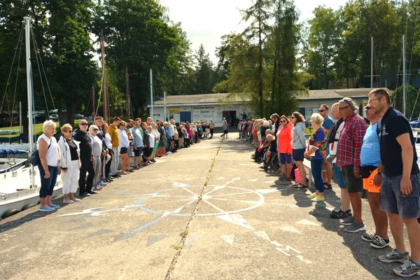 Zdjęcie pokazuje uczestników warsztatów żeglarskich na zbiórce przed ośrodkiem szkoleniowym na brzegu jeziora.